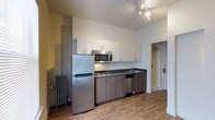 This single-floor space is thoughtfully designed to maximize functionality and comfort. The kitchen serves as a central highlight, featuring a full suite of stainless steel appliances including a Whirlpool refrigerator, over-the-range microwave, dishwasher, and a built-in cooktop. Sleek white upper cabinetry contrasts beautifully with the warm, wood-toned lower cabinets, while a dark countertop adds a contemporary finishing touch. A stainless steel sink sits neatly beneath the cabinetry, offering practical workspace. The kitchen is illuminated by modern track lighting mounted on the ceiling, ensuring ample brightness throughout. Warm wood-look flooring flows across the space, lending a cozy and inviting ambiance. A classic cast-iron radiator is visible along the wall, nodding to the building's character while providing efficient heating. A doorway off the kitchen leads to additional areas of the unit, offering a clear and logical layout. The overall design balances modern updates with timeless architectural details, creating a space that is both practical and visually appealing.