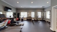 This single-level fitness center is thoughtfully designed to support a comprehensive workout experience. The space is finished with elegant crown molding and neutral gray walls, giving it a clean, polished aesthetic that balances functionality with style. Recessed lighting illuminates the room evenly throughout.

The cardio zone features multiple treadmills, elliptical trainers, and a recumbent bike, all positioned to face wall-mounted flat-screen televisions for an engaging workout experience. Large mirrors span entire wall sections, enhancing the sense of openness and allowing users to monitor their form. Natural light filters in through multiple windows fitted with white blinds, adding brightness to the space.

The strength training area is well-stocked with a multi-functional cable machine, a dedicated free weight rack offering a full range of dumbbells, a flat workout bench, and additional resistance training equipment. Stability balls and resistance accessories round out the available equipment.

A built-in water fountain station provides convenient hydration, and a French door with glass panes connects the fitness center to an adjacent room, ensuring easy access. Rubber flooring throughout provides both safety and durability. Motivational posters and a bulletin board add an encouraging atmosphere, making this fitness center a complete and inviting destination for daily exercise.