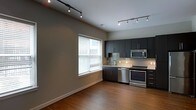 This single-floor space showcases a contemporary open-concept layout that seamlessly connects the living area and kitchen. The kitchen is outfitted with dark flat-panel cabinetry that extends to the upper walls, providing ample storage while contributing to the space's modern aesthetic. Stainless steel appliances, including a range and built-in microwave, complement the cabinetry, while a light-toned tile backsplash adds a subtle textural contrast. The countertops continue the clean, modern theme, and a single-basin sink is positioned beneath a window that allows natural light to fill the kitchen workspace.

Track lighting runs across the ceiling in both the living and kitchen areas, offering flexible, directed illumination throughout. The living area features warm hardwood flooring that extends seamlessly into the kitchen, creating a cohesive and inviting atmosphere. Neutral gray walls provide a versatile backdrop suitable for a variety of interior styles. A large window fitted with horizontal blinds in the living area allows for natural light while maintaining privacy. The overall ambiance is clean, minimalist, and functional.