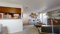 This single-floor residence offers a thoughtfully arranged layout that balances comfort and functionality throughout its main living areas. The carpeted living room serves as the heart of the home, featuring a textured ceiling, a built-in wet bar with warm wood cabinetry and granite-style countertops, glass display shelves, and a sliding glass door that opens to a private balcony, inviting natural light into the space. A dedicated dining area sits adjacent to the living room, illuminated by a classic chandelier and connected to the kitchen via a convenient pass-through window. The galley-style kitchen is efficiently designed with rich wood cabinetry lining both walls, granite-style countertops, wood-look flooring, and white appliances including a range and built-in microwave. The bedroom is a serene retreat featuring plush carpet, a ceiling fan, and a window that allows natural light to filter through sheer curtains. The full bathroom includes a wood-vanity with a granite-style countertop, a tub-shower combination with tile surround, and wood-look flooring. Throughout the residence, a warm, earthy aesthetic is maintained with consistent wood tones and neutral wall colors, creating a cohesive and welcoming ambiance.