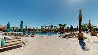This expansive outdoor resort-style complex offers a compelling array of amenities set against a striking desert mountain backdrop. At its centerpiece is a generously sized swimming pool with crystal-clear blue water, surrounded by wide, light-colored stone pool decking designed to accommodate rows of wooden lounge chairs and teal-accented umbrellas for ample sun and shade options.

Mature palm trees are planted throughout the grounds, providing a tropical atmosphere while complementing the desert landscape. A lazy river winds through the property, accessible via a dedicated pedestrian bridge complete with safety signage, stairways, and railings. The landscaping incorporates natural desert rock elements, adding to the cohesive Southwest aesthetic.

The complex includes a modern, multi-story clubhouse building featuring floor-to-ceiling glass curtain walls, colorful accent paneling in blue and yellow tones, and an outdoor bar area with a stone-faced counter and bar stool seating. Wide concrete staircases with built-in step lighting and steel railings connect the pool level to the upper terrace and clubhouse. Additional open concrete terrace space flanked by palm trees offers flexible use areas with panoramic views of the surrounding mountains and community. The overall design balances contemporary architecture with resort-inspired outdoor living.