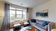 Located at 10 Kings Rd in Madison, NJ, this single-floor residence offers a thoughtfully designed layout with a cohesive contemporary aesthetic throughout. The living area is bright and welcoming, featuring large windows dressed with grommet-style curtains that frame views of the surrounding greenery and neighborhood. The space is furnished with a tufted gray sofa, mid-century accent chairs, and a warm wood coffee table, all anchored by a blue area rug. Ocean-inspired artwork adds a calming, unified tone to the room.

The kitchen is well-equipped with dark espresso cabinetry, stainless steel appliances including a gas range, over-the-range microwave, and refrigerator, complemented by light granite-style countertops and a tile backsplash. Rich hardwood-style flooring runs throughout the main living areas, lending warmth and continuity.

Both bedrooms are generously proportioned, each featuring natural light from well-placed windows, hardwood-style flooring, and neutral walls. One bedroom includes a workspace, while the other features a dark wood dresser with a decorative mirror. The bathroom is clean and modern, with white subway tile surrounding the tub-shower combo, a dark vanity with a white countertop, and light tile flooring.
