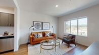 Located at 17100 SE Sunnyside Rd in Happy Valley, Oregon, this thoughtfully designed single-floor residence combines modern aesthetics with functional living. The open-concept layout seamlessly connects the kitchen, dining, and living areas, creating a cohesive and inviting atmosphere throughout.

The kitchen is a standout feature, outfitted with sleek gray cabinetry, stainless steel appliances including a range, microwave, dishwasher, and refrigerator, complemented by a crisp white subway tile backsplash and light countertops. Warm wood-tone flooring runs throughout the main living areas, adding natural warmth to the contemporary design.

The living room is bright and airy, benefiting from a large sliding window that frames views of the surrounding neighborhood and rolling Oregon hillside. The space accommodates comfortable seating and accent furnishings with ease.

The bedroom is carpeted for comfort and features its own window with outdoor views, providing natural light throughout the day. A dedicated closet area with hanging storage adds practical functionality. The overall color palette of soft whites and warm neutrals lends the space a clean, modern feel that is both welcoming and versatile.