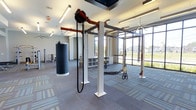 This contemporary fitness facility is designed to support a wide range of training styles within a spacious, open-plan layout. The space is anchored by a robust multi-functional training rig system, which supports suspension trainers, battle ropes, and pulley attachments — catering to both strength and conditioning workouts. A heavy punching bag hangs prominently, adding a dedicated space for combat-style training.

The floor is finished with durable, patterned carpet tiles in a neutral gray palette, providing comfort and traction underfoot. Overhead, the ceiling features recessed linear lighting that ensures consistent, well-distributed illumination throughout the space. A ceiling fan adds to the ventilation and comfort of the facility.

Large floor-to-ceiling windows along one wall flood the interior with abundant natural light while offering open views of the surrounding landscape, creating an energizing and welcoming atmosphere. Built-in storage cubbies are conveniently integrated along the wall, offering organized space for equipment and personal items.

Additional strength training equipment is visible in the background, suggesting the facility extends into further dedicated workout zones. The overall design balances functionality with a clean, modern aesthetic, making it a versatile and motivating environment for a variety of fitness activities.