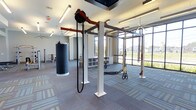 This contemporary fitness facility is designed to support a wide range of training styles within a spacious, open-plan layout. The space is anchored by a robust multi-functional training rig system, which supports suspension trainers, battle ropes, and pulley attachments — catering to both strength and conditioning workouts. A heavy punching bag hangs prominently, adding a dedicated space for combat-style training.

The floor is finished with durable, patterned carpet tiles in a neutral gray palette, providing comfort and traction underfoot. Overhead, the ceiling features recessed linear lighting that ensures consistent, well-distributed illumination throughout the space. A ceiling fan adds to the ventilation and comfort of the facility.

Large floor-to-ceiling windows along one wall flood the interior with abundant natural light while offering open views of the surrounding landscape, creating an energizing and welcoming atmosphere. Built-in storage cubbies are conveniently integrated along the wall, offering organized space for equipment and personal items.

Additional strength training equipment is visible in the background, suggesting the facility extends into further dedicated workout zones. The overall design balances functionality with a clean, modern aesthetic, making it a versatile and motivating environment for a variety of fitness activities.