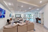 This single-floor community amenity space offers a generous, open layout designed for both relaxation and social gatherings. The kitchen area serves as a centerpiece, anchored by a stunning large-format marble island with seating for multiple guests, illuminated by a cluster of modern globe pendant lights. Adjacent cabinetry in a soft gray tone provides ample counter space and storage, complemented by stainless steel appliances, an undermount sink, and a shimmering herringbone tile accent wall. A vending machine adds convenient snack and beverage access.

The living room area is spacious and inviting, featuring multiple sectional sofas arranged around a dark coffee table atop a geometric blue and white area rug. Two large wall-mounted flat-screen TVs serve as focal points against a wainscoted white accent wall. Tray ceilings with recessed lighting throughout the space lend architectural elegance and a sense of added height.

A dedicated game zone includes a full-size pool table set on a textured area rug, while high-top bistro tables with cushioned bar-height chairs are positioned throughout for casual seating. Light wood-look tile flooring runs seamlessly across the entire space, tying together the clean, contemporary aesthetic with a warm and welcoming ambiance ideal for community use.