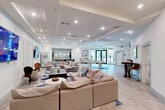 This single-floor community amenity space offers a generous, open layout designed for both relaxation and social gatherings. The kitchen area serves as a centerpiece, anchored by a stunning large-format marble island with seating for multiple guests, illuminated by a cluster of modern globe pendant lights. Adjacent cabinetry in a soft gray tone provides ample counter space and storage, complemented by stainless steel appliances, an undermount sink, and a shimmering herringbone tile accent wall. A vending machine adds convenient snack and beverage access.

The living room area is spacious and inviting, featuring multiple sectional sofas arranged around a dark coffee table atop a geometric blue and white area rug. Two large wall-mounted flat-screen TVs serve as focal points against a wainscoted white accent wall. Tray ceilings with recessed lighting throughout the space lend architectural elegance and a sense of added height.

A dedicated game zone includes a full-size pool table set on a textured area rug, while high-top bistro tables with cushioned bar-height chairs are positioned throughout for casual seating. Light wood-look tile flooring runs seamlessly across the entire space, tying together the clean, contemporary aesthetic with a warm and welcoming ambiance ideal for community use.