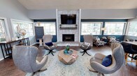 This single-story clubhouse is designed to serve as a welcoming communal gathering space, combining thoughtful architectural details with a carefully curated interior aesthetic. The main lounge area is anchored by a floor-to-ceiling stone fireplace with a built-in TV mount and rustic wood mantel, surrounded by swivel accent chairs and a geometric wood coffee table set atop a light area rug. Rich navy and teal accent walls contrast with light gray tones throughout, creating a vibrant yet cohesive color palette. Vaulted ceilings rise dramatically in portions of the space, enhanced by a modern multi-bulb chandelier that adds an industrial-chic touch. Abundant natural light pours in through multiple white-trimmed windows, offering views of the surrounding greenery. A separate seating nook features patterned armchairs and floor lamps for a more intimate setting. A kitchenette with a wood-topped bar counter, bar stools, stone tile backsplash, and appliances including a coffee station and refrigerator provides convenient amenities. A secondary open area with soaring double-height ceilings, arched decorative windows, and a dining table rounds out the space with additional versatility.