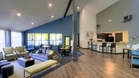 Located at 3201 Walnut Creek Dr in Raleigh, NC, this single-floor community clubhouse offers an expansive, open-concept gathering space designed for both relaxation and socializing. The interior is immediately striking thanks to its soaring vaulted ceilings, anchored by bold exposed beams and a prominent structural column that adds an industrial-chic character to the space.

The layout is thoughtfully organized into distinct zones that flow seamlessly into one another. A comfortable lounge area features plush seating, including a large sectional sofa accented with yellow throw pillows that echo the room's vibrant color palette. Adjacent to the lounge, a casual dining area is furnished with round tables and chairs, ideal for small group gatherings. The bold blue accent wall serves as a unifying backdrop, complemented by bright yellow window insets that bring energy and visual interest to the space.

Toward the rear, a sleek kitchen bar with dark cabinetry, stainless steel appliances, and counter-height seating provides a functional and stylish amenity hub. Recessed lighting throughout ensures the space is well-lit, while durable wood-look flooring and area rugs help define each zone. The overall ambiance is modern, welcoming, and well-appointed for community use.
