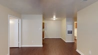 This single-floor space offers a straightforward, functional layout with an open living area that flows naturally toward the kitchen and bathroom. The flooring throughout is a warm-toned wood-style laminate, providing a cohesive look across the main living zone. Walls are finished in a soft neutral tone, complemented by white baseboards that add clean definition to each room.

The living area is open and unobstructed, allowing for flexible furniture arrangements. A flush-mount ceiling light provides overhead illumination, and the textured ceiling adds subtle visual interest. A partial wall divider separates the living space from the kitchen area, where warm wood-toned cabinetry is visible, offering practical storage. The kitchen appears to include countertop space along the cabinetry run.

Toward the rear of the space, a utility panel or HVAC access unit is mounted on the wall, indicating practical infrastructure within the unit. A glimpse into the bathroom reveals light-colored fixtures and tile, consistent with the overall neutral palette of the space. The layout is compact yet efficiently organized, making good use of the available floor plan on a single level.