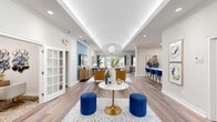 Located at 300 Industrial Pkwy in Saraland, Alabama, this single-story community clubhouse and leasing space presents a thoughtfully designed interior that balances professionalism with inviting comfort. The open-concept layout is anchored by wide-plank wood-look flooring that flows seamlessly throughout, unifying each functional area under one cohesive aesthetic.

The main gathering area commands attention with its tray ceiling featuring integrated cove lighting, which casts a soft ambient glow across the room. A central arched ceiling detail adds architectural interest, while recessed lighting supplements the natural light admitted through multiple large windows and glass-paned French doors. The seating arrangement incorporates a neutral sofa, richly toned mustard-gold accent chairs, a gold-pedestal coffee table, and royal blue ottomans, creating a welcoming lounge atmosphere punctuated by bold color.

A teal accent wall serves as a focal point for a wall-mounted flat-screen television positioned above a warm wood media console. Adjacent to the lounge, a bar-height counter with deep blue seating provides an additional gathering area. White-framed French doors with divided glass panes lead to private leasing offices, offering both transparency and separation. Natural light pours through the exterior glass doors, maintaining a bright and airy ambiance throughout the entire space.