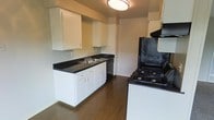 This single-story space includes a galley-style kitchen that serves as a central feature of the unit. The kitchen is designed for efficient use of space, with white cabinetry lining the walls and providing ample storage through both upper and lower cabinets. Dark countertops offer a contrasting aesthetic against the white cabinets, creating a classic two-tone look. A double-basin sink is conveniently positioned along the counter, accompanied by a built-in dishwasher. On the opposing side, a black gas range with multiple burners and an overhead ventilation hood is ready for everyday cooking. A flush-mount ceiling light illuminates the kitchen, ensuring a well-lit workspace. The flooring throughout is a dark hardwood-style material that adds warmth and cohesion to the overall design. The kitchen opens toward a hallway, suggesting a connected layout that flows into additional areas of the space. The overall ambiance is practical and straightforward, with a clean, no-frills design that prioritizes functionality and usability.