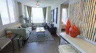 This single-floor residence showcases a thoughtfully designed open layout that seamlessly connects the living area to adjacent spaces. The living room serves as the focal point, featuring rich, dark hardwood flooring that extends throughout and provides a warm, grounding contrast to the lighter walls and furnishings. A ceiling fan with a light fixture anchors the room from above, complementing the natural light that pours in through large windows dressed with patterned green drapes. The seating arrangement includes a comfortable sofa paired with two contemporary accent chairs, centered around a sleek glass coffee table atop a textured area rug. A dark console unit along one wall provides storage and display space, adorned with decorative greenery and sculptural accents. Bold red ceramic vases on a console near the entryway add a striking pop of color. A glimpse through an open doorway reveals a bedroom beyond, suggesting a well-connected floor plan. The textured ceiling adds depth overhead, while the overall aesthetic balances modern design with warm, livable comfort.