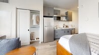Located at 2132 Stokes Blvd in Cleveland's vibrant University Circle neighborhood, this efficiently designed studio offers a thoughtfully organized single-floor layout that maximizes every square foot. The open-plan living space seamlessly integrates the sleeping, dining, and living areas, anchored by large floor-to-ceiling windows that flood the room with natural light and offer views of the surrounding cityscape. Warm wood-toned furnishings, including a built-in shelving unit with an integrated flat-screen TV, complement the light hardwood-style flooring throughout.

The kitchenette is well-appointed with gray cabinetry, white quartz countertops, a stainless steel refrigerator, a microwave, a sink with a matte black faucet, and white tile backsplash. Adjacent to the kitchen, a stacked in-unit washer and dryer provide added convenience. A sliding-door closet offers organized storage with hanging rods and shelving.

The bathroom features a clean, modern aesthetic with a gray vanity, white countertop, undermount sink, matte black fixtures, a large mirror, and a glass-enclosed shower and tub combination. Throughout the unit, a neutral white and gray palette creates a calm, contemporary ambiance ideal for comfortable urban living.