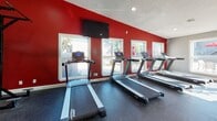 This fitness center is organized into several distinct workout zones, each designed to support a comprehensive exercise routine. The cardio area is lined with a row of modern treadmills positioned along a bold red accent wall, beneath a wall-mounted flat-screen television. A series of windows along this wall allow natural light to filter in while offering views of the outdoor surroundings, creating an energizing atmosphere for cardio training.

Adjacent to the treadmill area, a dedicated cycling and elliptical zone houses spin bikes and stair climbers on durable dark rubber flooring. Large windows in this section further enhance the sense of openness and connectivity to the outdoors. Recessed lighting in the textured ceiling provides consistent illumination throughout.

A separate area accommodates recumbent stationary bikes, set within a quieter section of the facility featuring neutral gray walls and high ceilings that contribute to an open, airy feel. Mirrored walls are incorporated into the strength training zone, which is equipped with weight machines, a power rack, free weights, and benches with red upholstery. Recessed lighting throughout the strength area ensures clear visibility, while the mirrored walls visually expand the space and allow users to monitor their form effectively.