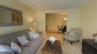This single-floor residence offers a thoughtfully arranged living space that balances comfort and functionality. The living room serves as the heart of the home, featuring soft, neutral wall tones and plush carpeting that lend a warm and relaxed feel. A stylish sofa paired with a patterned accent chair frames a dark wood coffee table, creating an inviting area for relaxation and conversation. A classic table lamp adds a soft, ambient glow that complements the overall warmth of the room. An open passageway connects the living room seamlessly to the dining area, promoting an airy, open-concept flow. The dining space is appointed with a dark-toned table and chairs, illuminated by an elegant chandelier overhead. Wall art adds a decorative touch, contributing to the cohesive and tasteful aesthetic of the space. Natural and artificial lighting work together throughout the interior to create a bright, welcoming ambiance. The continuity of carpeting and neutral color palette across both spaces ties the layout together harmoniously, making this residence feel both polished and livable.