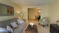 This single-floor residence offers a thoughtfully arranged living space that balances comfort and functionality. The living room serves as the heart of the home, featuring soft, neutral wall tones and plush carpeting that lend a warm and relaxed feel. A stylish sofa paired with a patterned accent chair frames a dark wood coffee table, creating an inviting area for relaxation and conversation. A classic table lamp adds a soft, ambient glow that complements the overall warmth of the room. An open passageway connects the living room seamlessly to the dining area, promoting an airy, open-concept flow. The dining space is appointed with a dark-toned table and chairs, illuminated by an elegant chandelier overhead. Wall art adds a decorative touch, contributing to the cohesive and tasteful aesthetic of the space. Natural and artificial lighting work together throughout the interior to create a bright, welcoming ambiance. The continuity of carpeting and neutral color palette across both spaces ties the layout together harmoniously, making this residence feel both polished and livable.