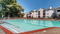 Located at 345 Buckland Hills Dr in Manchester, Connecticut, this outdoor amenity area centers around a generously sized lap pool with clearly marked swim lanes and crisp, aqua-blue water. The pool is framed by a expansive brick paver deck that provides ample space for lounging, with a variety of seating options including sun loungers, chairs, and umbrella-shaded tables arranged throughout the area. A classic white-sided clubhouse with a pitched roof and decorative dormers anchors one end of the pool deck, serving as a focal point and functional facility hub for the community. The entire pool area is enclosed by a black wrought iron fence, ensuring both safety and a polished aesthetic. Mature deciduous trees surround the perimeter, offering natural shade and a lush, park-like backdrop that enhances the sense of privacy and tranquility. The multi-story residential buildings of the community are visible nearby, featuring traditional New England architectural styling with white siding, pitched rooflines, chimneys, and private balconies. The overall ambiance is well-maintained and inviting, combining classic architectural design with thoughtfully arranged outdoor living spaces ideal for relaxation and recreation.