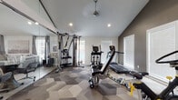 This fitness center is a comprehensively outfitted facility designed to support a wide range of workout routines. The space is unified by a striking geometric hexagonal carpet in varying shades of grey, which adds visual interest throughout. Vaulted ceilings with recessed lighting create an open, airy atmosphere, while multiple windows with white blinds allow natural light to fill the room from multiple angles.

The cardio section is well-stocked, featuring treadmills, elliptical trainers, and LeMond cycling bikes positioned in front of a wall-mounted flat-screen TV with an interactive fitness display, enabling on-demand workout classes. A multi-station cable machine with TRX suspension straps anchors the strength training area, complemented by a rack stocked with medicine balls, kettlebells, and stability balls of various sizes.

A full floor-to-ceiling mirrored wall runs along one side, visually expanding the space and providing form-checking capability during workouts. An open stretching and functional training zone offers ample room for floor exercises, plyo boxes, and other bodyweight movements. The facility is accessed through glass entry doors and includes a small administrative or reception area at the entrance, complete with a paper towel dispenser and waste bin for convenience.