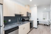 This thoughtfully designed single-floor studio offers a compact yet functional layout that makes efficient use of every square foot. The open corridor-style floor plan seamlessly connects the kitchen to the living and sleeping areas, creating a cohesive flow throughout the space.

The kitchen is outfitted with light wood cabinetry, stone-look countertops, a slate-tiled backsplash, a stainless steel sink, and a full suite of stainless steel appliances including a dishwasher, microwave, and refrigerator — providing everything needed for everyday living.

The sleeping area is bright and inviting, benefiting from a large window that allows natural light to fill the room. It includes a desk and chair, making it suitable for both rest and work or study. A wall-mounted flat-screen television adds a modern convenience. An additional sleeping nook provides extra flexibility, furnished with a bed and a small sofa, offering a versatile secondary space.

Light wood-toned flooring runs throughout, complementing the clean, white walls and recessed lighting that give the unit a contemporary, airy feel. A bold abstract painting in the hallway adds a striking artistic touch.

The bathroom is clean and well-appointed, featuring a combination tub and shower with glass sliding doors, a vanity with stone-look countertop, and a decorative woven mirror that adds warmth to the space.