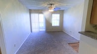 This single-floor residence offers a comfortable and functional layout designed for everyday living. The main living area is a spacious, open room finished with wall-to-wall carpeting in a neutral tone, providing a soft and inviting atmosphere underfoot. A ceiling fan with an integrated light fixture is centrally mounted on the textured ceiling, offering both air circulation and ambient lighting throughout the space.

Natural light flows generously into the room through two key openings: a large sliding glass door dressed with vertical blinds, and a traditional window positioned on the adjacent wall. Together, these features create a bright, airy environment that feels open and welcoming. The sliding door suggests access to an outdoor area such as a patio or balcony, further enhancing the sense of connection to the exterior.

The walls are finished in a clean, light neutral tone, complemented by white baseboards that frame the room with a crisp, tidy appearance. A partial view of an adjacent area with hard flooring is visible, hinting at a seamlessly connected secondary space. Overall, the residence presents a clean, move-in ready environment with practical finishes and an abundance of natural light.