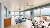 This rooftop entertainment space combines a bright, enclosed sunroom with a sprawling open-air deck, offering a versatile setting designed for leisure and social gatherings. The interior sunroom is defined by teal-painted walls, light tile flooring, and an abundance of natural light streaming through large sliding glass doors and oversized windows that frame panoramic views of the surrounding cityscape and open water. Ceiling fans keep the space comfortable year-round, and the room is well-suited for recreation, featuring areas for ping pong, air hockey, and table games such as checkers. A dedicated dining nook with a glass-top table and chairs provides a relaxed spot to take in the views. The seamless transition between indoor and outdoor areas is a standout feature — sliding glass doors open directly onto a generously sized wood-plank deck that wraps around the sunroom. The deck is outfitted with built-in wood bench seating and planter boxes, creating defined gathering zones. A railing system along the perimeter ensures safety while preserving unobstructed sightlines to the urban skyline and waterfront beyond. Together, the indoor and outdoor areas deliver a compelling rooftop retreat with exceptional elevated views.