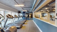 Located at 5290 Big Island Dr in Jacksonville, FL, this expansive fitness center is designed to accommodate a wide range of workout needs within a single, thoughtfully organized floor. The space features an industrial aesthetic defined by exposed concrete ceilings with visible ductwork and piping, complemented by a two-tone blue and white wall treatment that lends the facility a modern, professional character.

The main exercise floor is divided into distinct functional zones connected by a curving teal-colored pathway set into neutral patterned carpet tiles. A dedicated cardio section lines the perimeter windows, housing multiple Precor treadmills and ellipticals that offer outdoor views of surrounding palm-lined walkways and balconies, allowing natural light to fill the area. Mounted televisions are positioned throughout for convenience during workouts.

The strength training area features an extensive selection of Precor cable machines, selectorized equipment, and free-standing strength stations, all arranged alongside large wall mirrors that visually expand the space. Stability balls and functional training accessories are also available throughout.

A separate studio area provides a quieter zone outfitted with spin bikes, a WaterRower, and open floor space for stretching or group activities. Locker room access and a water fountain are conveniently located nearby, completing this well-rounded facility.