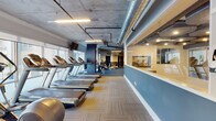 Located at 5290 Big Island Dr in Jacksonville, FL, this expansive fitness center is designed to accommodate a wide range of workout needs within a single, thoughtfully organized floor. The space features an industrial aesthetic defined by exposed concrete ceilings with visible ductwork and piping, complemented by a two-tone blue and white wall treatment that lends the facility a modern, professional character.

The main exercise floor is divided into distinct functional zones connected by a curving teal-colored pathway set into neutral patterned carpet tiles. A dedicated cardio section lines the perimeter windows, housing multiple Precor treadmills and ellipticals that offer outdoor views of surrounding palm-lined walkways and balconies, allowing natural light to fill the area. Mounted televisions are positioned throughout for convenience during workouts.

The strength training area features an extensive selection of Precor cable machines, selectorized equipment, and free-standing strength stations, all arranged alongside large wall mirrors that visually expand the space. Stability balls and functional training accessories are also available throughout.

A separate studio area provides a quieter zone outfitted with spin bikes, a WaterRower, and open floor space for stretching or group activities. Locker room access and a water fountain are conveniently located nearby, completing this well-rounded facility.