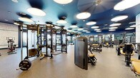 Located at 1010 E Orange St in Tempe, Arizona, this fitness center offers a generously sized, single-floor workout facility designed to accommodate a wide range of training needs. The space is defined by its bold aesthetic: a deep navy blue ceiling adorned with large circular pendant lights creates a modern, energetic atmosphere throughout.

The facility is organized into distinct functional zones. The strength training area is well-equipped with a comprehensive selection of Life Fitness selectorized machines, multi-functional cable systems with TRX attachments, and power racks. Floor-to-ceiling mirrors line the walls of the free weights section, which features a full rack of dumbbells, adjustable benches, and barbells with color-coded Olympic weight plates.

The cardio zone is positioned near floor-to-ceiling windows that allow natural light to pour in and offer views of the surrounding Tempe landscape. It is outfitted with treadmills, ellipticals, stationary cycles, and air bikes. A dedicated open floor area with rubber matting provides space for functional training and stretching.

The facility features durable speckled rubber flooring throughout, a decorative mosaic-patterned accent wall, and built-in storage cubbies. The entrance is accessible via glass double doors with keycard access, reflecting the space's modern, amenity-grade design.