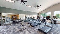 This fitness center is thoughtfully designed to support a comprehensive workout experience across multiple distinct zones. The main exercise area boasts impressively vaulted ceilings with large arched windows and expansive floor-to-ceiling glass panels that flood the space with natural light and offer views of the surrounding greenery. Patterned, textured carpet tiles cover the floors throughout, lending a modern yet functional aesthetic. Ceiling fans are strategically positioned overhead to maintain comfortable airflow during workouts. The cardio section is outfitted with a row of True-brand treadmills and elliptical machines, all positioned to take advantage of the natural light and outdoor views. A full rack of dumbbells and adjustable weight benches provide ample free weight options, while full-length mirrored walls amplify the sense of space and allow for form-checking during exercises. The strength training area features a variety of cable machines and resistance equipment, offering a well-rounded selection for muscle conditioning. A separate spin studio with light wood flooring and recessed lighting houses several stationary cycling bikes, creating a focused and dedicated space for cycling workouts. Connecting corridors lead to additional strength equipment, rounding out this facility as a complete, multi-functional fitness destination.