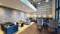Located at 1800 Indigo Square in Mount Pleasant, South Carolina, this thoughtfully designed lounge space blends industrial and contemporary aesthetics to create an inviting, upscale atmosphere. The room immediately draws the eye to a dramatic floor-to-ceiling built-in bookshelf wall, lined with neatly arranged volumes and softly illuminated by integrated lighting, creating both visual depth and a warm, scholarly ambiance.

Contrasting the clean lines of the shelving, a textured exposed brick wall adds a raw, industrial character to the space. Rich hardwood flooring runs throughout, grounding the room with warmth and continuity. A mezzanine-level railing with sleek black metal detailing is visible overhead, hinting at the vertical scale of the space and contributing to its open, airy feel.

The seating area is carefully curated with a mix of plush sofas, accent chairs, and low wooden coffee tables arranged to encourage both intimate conversation and relaxed lounging. Recessed ceiling lighting and scattered floor lamps provide layered illumination throughout. The overall design strikes a balance between comfort and sophistication, making it a refined and functional gathering space.