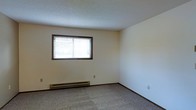 This single-floor space presents a straightforward and functional design. The room visible in the image features neutral-toned walls that create a clean, understated backdrop, complemented by wall-to-wall carpeting in a muted gray tone that adds a layer of comfort underfoot. A textured popcorn ceiling overhead gives the space a classic, traditional character.

A centrally placed window fitted with horizontal blinds allows for natural light while maintaining privacy. Beneath the window runs a baseboard heater, providing an efficient and unobtrusive heating solution positioned along the exterior wall. The room's corner angles are clean and well-defined, contributing to a tidy, orderly appearance.

The space is minimal in detail, with standard electrical outlets positioned along the walls, offering practical utility. The overall ambiance is quiet and neutral, making it a versatile blank canvas suitable for a variety of uses. The single-floor layout ensures easy accessibility throughout, and the understated design elements suggest a space that prioritizes function and simplicity.