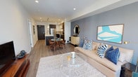 This thoughtfully designed single-floor residence combines modern aesthetics with functional living. The open-concept layout seamlessly connects the living, dining, and kitchen areas, creating a cohesive and inviting atmosphere. The living room features warm wood-look flooring, a muted blue accent wall, recessed lighting, and curated décor including bold artwork and a plush sectional sofa. A round glass coffee table and area rug anchor the space with understated elegance.

The kitchen is a standout feature, outfitted with two-tone dark and white cabinetry, white quartz countertops, stainless steel appliances including a gas range and French door refrigerator, and a center island with a undermount sink and pendant lighting above. The adjoining dining area accommodates a round glass table surrounded by mid-century modern chairs.

The primary bedroom offers a calming retreat with a cool-toned accent wall, recessed lighting, plush carpeting, and natural light from a pair of windows. It is complemented by a spacious walk-in closet with multi-level shelving and hanging rods. A second bedroom features similar clean, bright finishes. The bathroom is finished with large-format tile flooring, a dark-cabinetry vanity with a white countertop, and a full-width mirror, exuding a clean, contemporary style throughout.
