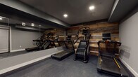 This dedicated fitness facility is thoughtfully designed to deliver a complete workout experience within a compact, purposeful layout. The space is anchored by a striking reclaimed wood accent wall that adds warmth and visual character against the predominantly dark, charcoal-toned interior. Durable rubber flooring in a dark speckled finish covers the entire area, providing traction and impact absorption ideal for athletic use.

The cardio zone houses multiple treadmills and a stair-climbing machine, all positioned along the accent wall and reflected in a large floor-to-ceiling mirror that spans a full wall, enhancing the sense of depth and allowing users to monitor their form. Recessed ceiling lighting provides bright, even illumination throughout.

A dedicated functional training area features a well-organized multi-tier equipment rack stocked with kettlebells, medicine balls, resistance tools, and stability balls, offering a full range of strength and conditioning options. Open floor space adjacent to the rack supports stretching, bodyweight exercises, and dynamic movements.

Entry to the fitness space is through sleek, full-glass double doors with dark metal framing, labeled for aerobics use, suggesting this facility is part of a larger multi-use building. The overall design blends industrial and rustic elements to create a motivating, professional-grade environment.