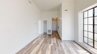 Located at 507 Grand St in Trenton, NJ, this thoughtfully designed one-bedroom, two-bathroom residence offers a clean, modern aesthetic throughout its well-appointed single floor. The entry hallway welcomes you with light wood-tone flooring, crisp white walls, and a decorative gold flush-mount light fixture. An elegant arched window at the end of the corridor floods the space with natural light, hinting at the building's distinctive architectural character.

The spacious living room features soaring ceilings that lend an open, airy feel, complemented by the same light hardwood-style flooring found throughout the unit. The generously sized primary bedroom includes a double-door closet and maintains the home's consistent neutral palette. The kitchen is outfitted with white cabinetry, dark stone countertops, a stainless steel refrigerator, and an electric range, all set atop striking marble-look tile flooring. A tall gridded window provides natural light and ventilation. Both bathrooms are finished in clean white tones, with one featuring a full tub-shower combination.

A standout feature of this residence is access to a large rooftop deck, finished with wood-tile decking and privacy fencing. The rooftop showcases the building's signature arched windows and offers sweeping open-air views of the surrounding Trenton skyline and treetops.