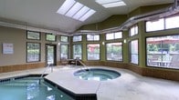 This single-story indoor aquatic space is designed to provide a bright, resort-like environment for relaxation and recreation. The room's most striking architectural feature is its vaulted ceiling, which incorporates multiple large skylights that flood the space with natural light throughout the day. Exposed industrial-style ductwork runs along the upper portion of the ceiling, adding a functional yet visually distinctive element to the design.

The walls are finished in a warm olive green and lined with an expansive array of multi-pane windows positioned at both mid and upper levels, offering views of an outdoor deck and surrounding greenery. This generous glazing ensures the interior remains bright and connected to the outdoors. Tile wainscoting lines the lower walls, complementing the neutral stone-toned pool deck.

The aquatic area includes an indoor swimming pool along one edge and a centrally positioned circular hot tub, finished with dark tile surround and accessible via metal handrails. A small seating area with a table and chairs is conveniently located poolside, providing a comfortable spot to relax. The overall ambiance is open, airy, and well-appointed.
