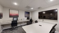 Located at 211 Warren Ave in Baltimore, MD, this single-floor business center offers a well-appointed workspace designed to support both collaborative and individual work needs. The space is accessed through glass-paneled doors that lend a professional, open feel to the entry corridor, which features neutral carpet and clean white walls.

The interior is divided into two functional zones. A dedicated workstation area runs along a long granite-topped counter, equipped with desktop computers, a printer, and ergonomic task chairs — providing ample space for individual productivity. Above the counter, bold framed artwork adds visual interest against the neutral walls.

Adjacent to the workstation area is a private conference room, distinguished by a striking mosaic-patterned accent wall in deep blue and white tones. A large white rectangular table anchors the room, surrounded by black mesh chairs, while a wall-mounted flat-screen display facilitates presentations and meetings. A second flat-screen display is also available in the main workstation area.

Throughout the space, a drop ceiling with recessed lighting provides consistent, professional illumination. Patterned carpet tile flooring in grey tones ties the areas together cohesively. The overall ambiance is sleek, modern, and functional, reflecting a well-designed professional environment suited for business use.