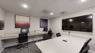 Located at 211 Warren Ave in Baltimore, MD, this single-floor business center offers a well-appointed workspace designed to support both collaborative and individual work needs. The space is accessed through glass-paneled doors that lend a professional, open feel to the entry corridor, which features neutral carpet and clean white walls.

The interior is divided into two functional zones. A dedicated workstation area runs along a long granite-topped counter, equipped with desktop computers, a printer, and ergonomic task chairs — providing ample space for individual productivity. Above the counter, bold framed artwork adds visual interest against the neutral walls.

Adjacent to the workstation area is a private conference room, distinguished by a striking mosaic-patterned accent wall in deep blue and white tones. A large white rectangular table anchors the room, surrounded by black mesh chairs, while a wall-mounted flat-screen display facilitates presentations and meetings. A second flat-screen display is also available in the main workstation area.

Throughout the space, a drop ceiling with recessed lighting provides consistent, professional illumination. Patterned carpet tile flooring in grey tones ties the areas together cohesively. The overall ambiance is sleek, modern, and functional, reflecting a well-designed professional environment suited for business use.