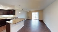 This single-story, 1-bedroom, 1-bathroom unit offers a thoughtfully designed open-concept layout that maximizes its functional living space. The main living and dining area is highlighted by rich hardwood-style flooring, recessed lighting, and a sliding glass door that fills the space with natural light while providing direct access to a private balcony with outdoor views.

The kitchen is seamlessly integrated into the living area via a peninsula with granite-style countertops and a pendant light overhead. Dark espresso cabinetry lines the walls, complemented by matching black appliances, including a range, microwave, and dishwasher, alongside a stainless steel sink. The cohesive design creates a polished, functional workspace.

The bedroom is a comfortable retreat featuring plush carpeting, neutral warm-toned walls, and a window with blinds that allows for natural light. A closet is accessible nearby, providing practical storage.

The bathroom features dark espresso cabinetry with a granite-style vanity countertop, a large mirror with overhead lighting, hardwood-style flooring, and a walk-in shower area. The overall ambiance throughout the unit is clean, modern, and move-in ready.
