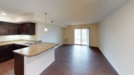 This single-story, 1-bedroom, 1-bathroom unit offers a thoughtfully designed open-concept layout that maximizes its functional living space. The main living and dining area is highlighted by rich hardwood-style flooring, recessed lighting, and a sliding glass door that fills the space with natural light while providing direct access to a private balcony with outdoor views.

The kitchen is seamlessly integrated into the living area via a peninsula with granite-style countertops and a pendant light overhead. Dark espresso cabinetry lines the walls, complemented by matching black appliances, including a range, microwave, and dishwasher, alongside a stainless steel sink. The cohesive design creates a polished, functional workspace.

The bedroom is a comfortable retreat featuring plush carpeting, neutral warm-toned walls, and a window with blinds that allows for natural light. A closet is accessible nearby, providing practical storage.

The bathroom features dark espresso cabinetry with a granite-style vanity countertop, a large mirror with overhead lighting, hardwood-style flooring, and a walk-in shower area. The overall ambiance throughout the unit is clean, modern, and move-in ready.