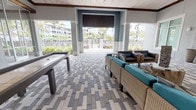 This single-story amenity pavilion is designed as a semi-open-air gathering space, blending indoor comfort with an outdoor ambiance. The structure features soaring ceilings clad in whitewashed wood planks, accented by dark exposed beams and ceiling fans that enhance air circulation throughout. The patterned paver flooring in varied shades of gray, beige, and white runs continuously across the entire space, reinforcing its cohesive coastal aesthetic.

The lounge area is anchored by wicker sectional sofas and armchairs with vibrant teal cushions, arranged around a textured stone accent wall mounted with a flat-screen television. A shuffleboard table and ping-pong table provide dedicated game areas within the pavilion. Retractable roller shades along the open sides offer flexible shade and privacy.

A fully equipped catering kitchen runs along one side of the pavilion, featuring granite-topped dark cabinetry, stainless steel appliances including a commercial range with overhead exhaust hood, and a decorative seashell-patterned tile backsplash. The open perimeter of the pavilion frames views of a gated pool area with palm trees and a multi-story residential building beyond, creating a bright, resort-inspired atmosphere throughout.