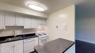 This single-floor space presents a well-organized and functional layout centered around a practical kitchen. The kitchen is outfitted with classic white cabinetry that provides ample storage, complemented by dark stone-finish countertops that offer a subtle contrast and generous prep space. White appliances, including a gas range and dishwasher, are seamlessly integrated beneath the cabinetry, contributing to a cohesive and tidy aesthetic. A range hood is positioned above the stove, ensuring proper ventilation during cooking. The kitchen also benefits from a peninsula or breakfast bar with matching dark countertop, providing additional workspace or casual seating. Overhead, a large oval flush-mount light fixture illuminates the space with bright, even lighting. The walls are finished in a clean, neutral tone that enhances the sense of openness and keeps the overall ambiance light and airy. Utility panels are neatly recessed into the wall, maintaining the clean lines of the space. The single-floor layout promotes easy accessibility and flow throughout, making this a practical and inviting space suited for everyday living.