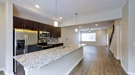 This two-storey space offers a thoughtfully designed open-concept main floor where the kitchen and living area flow seamlessly together. The kitchen is appointed with rich, dark espresso cabinetry that extends to the upper walls, providing generous storage. Stainless steel appliances, including a refrigerator, microwave, and range, complement the space's contemporary aesthetic. A striking granite-topped island serves as the kitchen's centerpiece, featuring an undermount sink and offering additional seating and prep space. A mosaic tile backsplash adds texture and visual interest behind the range area. Pendant lighting hangs above the island, reinforcing the modern design language, while recessed lighting provides consistent illumination throughout. The adjacent living area is open and airy, anchored by a ceiling fan and framed by a large window that draws in natural light. Durable hardwood-style flooring runs continuously through both the kitchen and living spaces, tying the areas together cohesively. Neutral grey wall tones complement the darker cabinetry and light granite surfaces, creating a balanced and inviting atmosphere. The second floor provides additional living space above the main level.