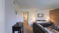 This single-floor space offers a comfortable and practical layout, with the living area serving as the central focus. The room features neutral-toned walls and carpet, creating a warm and inviting atmosphere. A dark upholstered sofa, accented with patterned and green throw pillows, anchors the seating area, while a matching dark console table adds a functional touch along one wall. Framed artwork decorates the walls, contributing to a lived-in, homey feel. A table lamp provides warm ambient lighting, complementing the natural light filtering into the space. The textured popcorn ceiling adds a classic character to the interior. A hallway visible from the living area connects to an adjacent bedroom, which features its own chandelier-style light fixture and warm décor, suggesting a well-connected and efficiently arranged floor plan. Ceiling vents throughout indicate a central HVAC system for year-round comfort. The overall ambiance is relaxed and approachable, with a straightforward design that emphasizes everyday functionality and comfort.