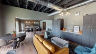 This single-floor community space showcases a striking industrial-modern aesthetic, defined by exposed black structural beams, large-diameter silver HVAC ductwork, and dark ceiling trusses that lend a loft-like character to the open-plan interior. Track lighting runs along the ceiling framework, providing ample illumination throughout.

The lounge area features warm hardwood flooring and is furnished with a tan leather sofa, teal accent chairs, and a coffee table set atop a patterned area rug. A bank of floor-to-ceiling grey cabinetry lines one wall, offering generous built-in storage. A contemporary globe chandelier adds a refined focal point overhead.

The fully equipped kitchen is a standout feature, boasting dark lower cabinetry, crisp white quartz countertops, a textured dark stone tile backsplash, open wood shelving, and a large central island with bar stools. Amber glass pendant lights hang above, complementing the exposed wood beam accents. The kitchen includes a stainless steel sink, dishwasher, and appliances.

A dining area with a long communal table and modern chairs connects the kitchen to the lounge. Toward the rear, large sliding glass doors and expansive windows flood the space with natural light and open directly to an outdoor patio, seating area, and pool — seamlessly blending indoor and outdoor living.