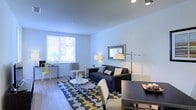 This single-floor space offers a well-designed, open-concept layout that seamlessly connects the living and dining areas into one cohesive environment. The living area is anchored by a dark sofa accented with decorative pillows, a bold geometric-patterned area rug in grey, yellow, and white tones, and a round white coffee table that adds a clean, modern touch. A wall-mounted mirror and a sleek arc floor lamp enhance the sense of depth and light in the room. The dining area sits adjacent to the living space, featuring a dark table paired with contemporary upholstered chairs, creating a functional yet stylish setting. A large window dressed with horizontal blinds floods the space with natural light, highlighting the smooth light grey walls and durable hardwood-style flooring that runs throughout. A compact desk and chair near the window provide a practical workspace without disrupting the open feel. Track lighting on the ceiling adds versatility to the overall illumination. The space strikes a balance between comfort and modern aesthetic, with a cohesive color palette of grey, white, and yellow accents tying all areas together in a fresh, inviting atmosphere.