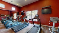 This fitness center offers a comprehensive workout environment designed to accommodate a full range of exercise needs. The space is defined by its bold, deep-red walls that create an energizing atmosphere, complemented by a multicolored patterned carpet and wood-look flooring that adds warmth throughout.

The cardio zone is well-appointed with multiple treadmills, elliptical trainers, and a stationary bike, each positioned to take advantage of large windows that frame views of the surrounding outdoor greenery. Wall-mounted flat-screen televisions are strategically placed throughout the space, providing entertainment during workouts.

The strength training area features a variety of cable machines, weight benches, and resistance equipment, offering users a thorough full-body training experience. Full-length mirrors line the walls, visually expanding the space and allowing for proper form monitoring during exercise.

Natural light flows in generously through both large lower windows and a row of clerestory windows positioned high along the upper walls, brightening the interior throughout the day. Recessed lighting and a ceiling fan further enhance comfort and visibility. The space also includes a set of French doors leading to an adjacent area, adding to its functionality. Overall, this fitness center provides a dynamic, motivating environment for a wide variety of workout routines.