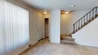 This three-floor residence offers a practical and well-organized layout designed for comfortable living. The entry level, pictured here, showcases neutral-toned wall-to-wall carpeting that lends warmth to the space. A built-in closet with a hanging rod provides convenient storage near the entryway, while a paneled white door with brass hardware serves as the main access point. Vertical blinds on the window allow for adjustable natural light control. Flush-mount ceiling light fixtures illuminate the area with a soft, warm glow, and floor vents indicate a central HVAC system throughout. A carpeted staircase with a dark metal railing connects this level to the upper floors, suggesting a thoughtfully designed vertical flow through the home. The walls are finished in a clean, neutral beige tone that complements the light-colored carpet, creating a cohesive and understated aesthetic throughout the visible areas. The open layout of this entry level offers flexibility in how the space can be utilized, with clear sightlines and a straightforward, functional design that carries through the rest of the home.