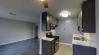 This single-floor space showcases a thoughtfully designed kitchen that serves as the focal point of the layout. The kitchen features dark, espresso-toned cabinetry that extends to the ceiling, providing generous storage capacity. Marble-style countertops add a touch of elegance, complementing the stainless steel appliances, which include a gas range, built-in microwave, and sink. The light wood-toned flooring in the kitchen transitions to darker hardwood-style flooring in the adjacent living area, visually distinguishing the two spaces while maintaining a cohesive aesthetic. The walls are finished in a cool, neutral gray tone throughout, contributing to a calm and modern ambiance. Flush-mount ceiling light fixtures provide consistent illumination, while a ceiling fan in the living area adds both functionality and airflow. The open sightline between the kitchen and living area creates an interconnected flow, making the space feel open and practical. A doorway visible in the background suggests additional rooms beyond, indicating a functional and well-organized floor plan overall.