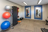 Located at 401 S Gallaher View Rd in Knoxville, Tennessee, this single-floor fitness center is thoughtfully designed to support a comprehensive workout experience. The facility is organized into distinct exercise zones, each equipped with purpose-specific equipment and connected by an open, functional layout. The cardio area features a row of Spirit treadmills positioned along large windows that offer direct views of the outdoor pool and surrounding wooded landscape, filling the space with natural light and scenic outdoor views. Mounted televisions are available throughout, providing entertainment during workouts. An adjacent cycling and elliptical zone houses multiple stationary bikes and an elliptical trainer, separated by a glass partition wall that maintains visual openness while defining the space. The free weights area is appointed with a full rack of dumbbells, a weight bench, and a full-length mirror set against a bold blue accent wall, creating a motivating environment for strength training. Additional accessories including stability balls and medicine balls are also available. The flooring throughout is durable carpet tile, and the textured ceilings are illuminated by fluorescent strip lighting. A louvered utility door is accessible from the main corridor. The overall ambiance is clean, modern, and functional, with a color palette of neutral grays accented by deep blue and orange details.
